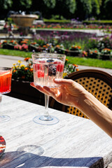 Hand holding berry cocktail with ice outdoors, Close-up of a hand holding a refreshing cocktail with ice, raspberries, and blackberries at an outdoor café garden in summer.