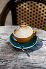 Cappuccino with latte art in yellow cup on cafe table, Freshly made cappuccino with creamy latte art served in a yellow cup on a turquoise saucer with spoon at an outdoor café.