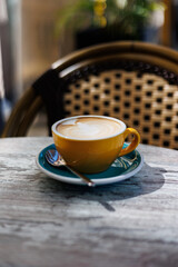 Cappuccino with latte art in yellow cup on cafe table, Freshly made cappuccino with creamy latte art served in a yellow cup on a turquoise saucer with spoon at an outdoor café.