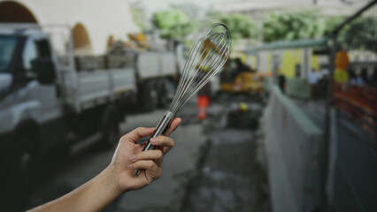 Hand holding whisk in front of a construction site with trucks on a busy street under a clear sky,...