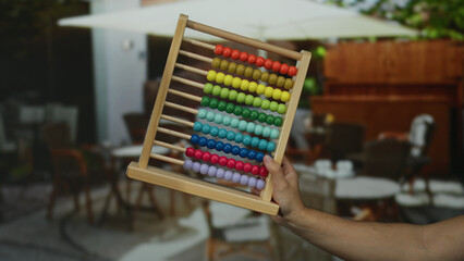 Man holding colorful abacus on outdoor restaurant terrace with blurred background showcasing tables and chairs, evoking a playful educational scene in a city setting.