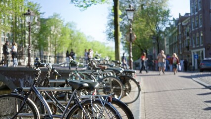 Bikes parked along a canal path in amsterdam with defocused people and trees in the sunny background creating a bokeh effect on the bustling street.