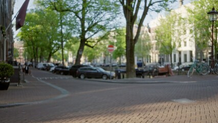 Blurred amsterdam street scene with defocused people, green trees, bicycles, and historic buildings creating a serene cityscape in the netherlands with a peaceful urban atmosphere.