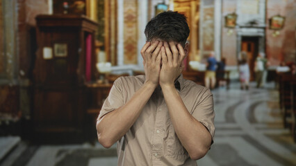 Young hispanic man standing in glasses covering face with both hands in ornate church building; shame.