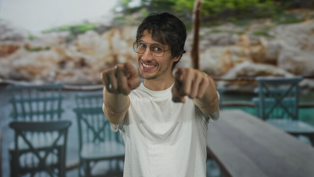 Man wearing glasses and white shirt smiling and pointing finger at camera on restaurant building terrace; invitation.