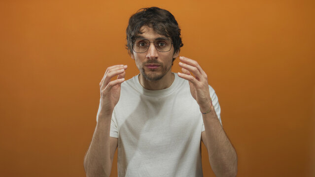 Man wearing glasses and white t shirt holds hands beside face in studio eyes fixed on camera; confusion doubt.
