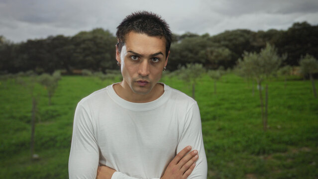 Young man with arms crossed standing in green park with trees and cloudy sky, wearing a white shirt and earrings, exuding a thoughtful expression in outdoor environment.
