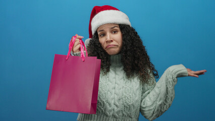 Woman wearing santa hat and sweater holds pink shopping bag with confused expression in front of blue background indicating holiday shopping dilemma.