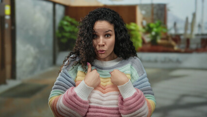 Woman in colorful sweater stands outdoors pointing at herself with a surprised expression on a...