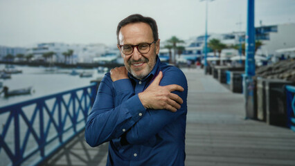 Middle-aged man in blue shirt embracing himself on a seaside promenade with boats and buildings in the background.