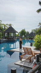 Woman walking by luxurious tropical resort pool surrounded by lush greenery and palm trees, embodying a serene holiday atmosphere.