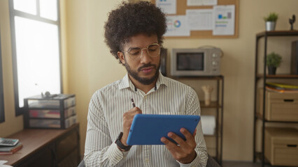 African american man using tablet in modern office with documents and plants visible indoors reflecting a focused work environment.