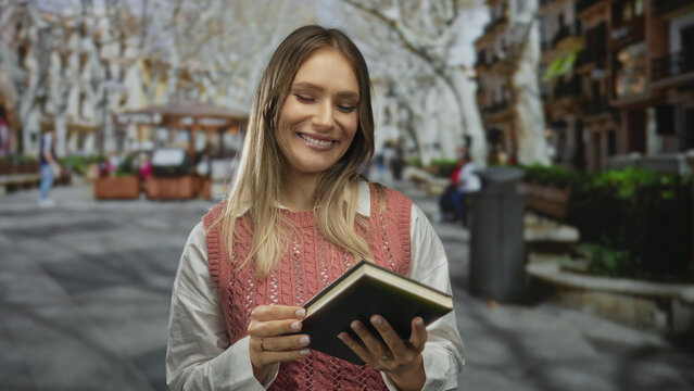 Woman reading book on city street, charming smile, sunny day, blonde hair, pink sweater, cheerful atmosphere, urban setting, outdoor scene, historic architecture.