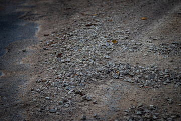 A detailed view of a weathered gravel and dirt road with uneven clusters of small gray stones scattered across the surface, featuring patches of exposed ground and a few yellow leaves