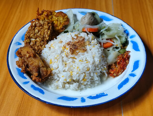 A portion of typical Indonesian mixed rice is served on a classic enamel plate with a blue and white pattern, consisting of corn rice, soup, fried tempeh, fried chicken, chili sauce, etc.