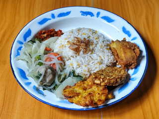A portion of typical Indonesian mixed rice is served on a classic enamel plate with a blue and white pattern, consisting of corn rice, soup, fried tempeh, fried chicken, chili sauce, etc.