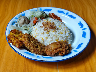 A portion of typical Indonesian mixed rice is served on a classic enamel plate with a blue and white pattern, consisting of corn rice, soup, fried tempeh, fried chicken, chili sauce, etc.