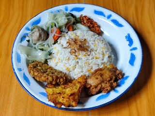 A portion of typical Indonesian mixed rice is served on a classic enamel plate with a blue and white pattern, consisting of corn rice, soup, fried tempeh, fried chicken, chili sauce, etc.