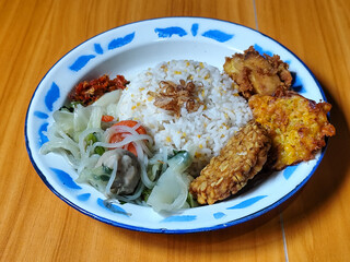 A portion of typical Indonesian mixed rice is served on a classic enamel plate with a blue and white pattern, consisting of corn rice, soup, fried tempeh, fried chicken, chili sauce, etc.