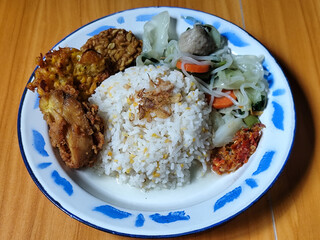 A portion of typical Indonesian mixed rice is served on a classic enamel plate with a blue and white pattern, consisting of corn rice, soup, fried tempeh, fried chicken, chili sauce, etc.