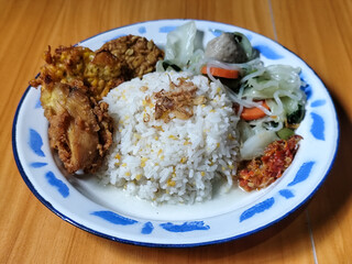 A portion of typical Indonesian mixed rice is served on a classic enamel plate with a blue and white pattern, consisting of corn rice, soup, fried tempeh, fried chicken, chili sauce, etc.
