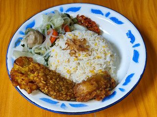 A portion of typical Indonesian mixed rice is served on a classic enamel plate with a blue and white pattern, consisting of corn rice, soup, fried tempeh, fried chicken, chili sauce, etc.