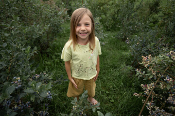 Among the green bushes in the garden, there is a girl wearing a yellow T-shirt with unicorns in yellow shorts, smiling broadly.