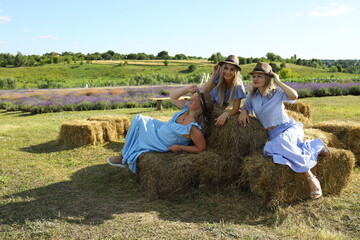 Three friends relax in a lavender field, sitting on hay bales while wearing summer dresses and hats. The bright sun illuminates the green landscape around them. © Rita