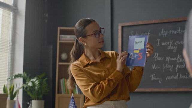 Medium shot of young Caucasian female teacher in glasses and mustard blouse pointing at book in her hands while addressing students during interactive classroom lesson