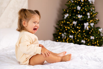 Fototapeta premium Sad and depressed toddler girl in casual bodysuit sits on a bed in front of a blurred Christmas tree background and crying. Childhood and emotions concept