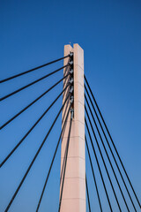 Obraz premium Close-up view of a modern cable-stayed bridge pylon with black suspension cables against a clear blue sky, showcasing contemporary civil engineering