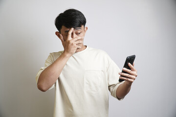 A shocked Asian man in a beige t-shirt covers his face with his hand while looking at his smartphone, reacting to something surprising or embarrassing.