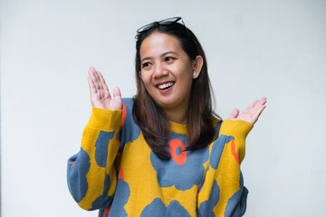 Young Asian woman in colorful sweater smiling while standing in front of a white wall, casual portrait concept.