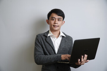  A confident young Asian businessman in a grey suit and open collar shirt holds a laptop, smiling while looking directly at the camera.