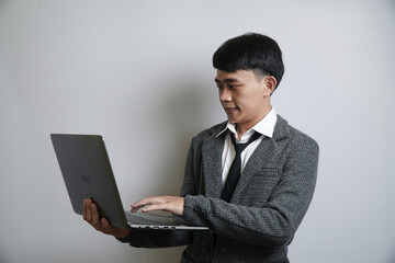 A focused young Asian businessman in a grey suit and tie is using a laptop, with a serious and concentrated expression.