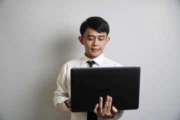 Young Asian man in formal clothes looking at laptop screen with focus and slight smile, standing against neutral background.