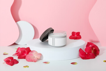Cosmetic jar mockup with a blank label for branding. Glass cream container on a white podium, decorated with red hydrangea flowers against a modern pink background.