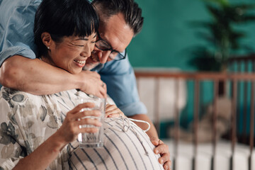 Happy pregnant woman holding glass of water and embracing husband in nursery