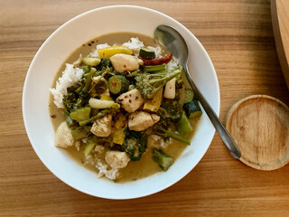 Chicken Thai green curry with vegetables broccoli, served with rice sprinkled with chili in a white bowl with silver spoon, on a wooden worktop, Asian cuisine and flavorful food presentation