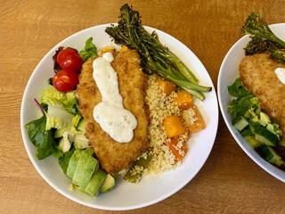 Dinner of breaded chicken, fresh salad, couscous, and broccoli served in a white bowl on a wooden worktop, highlighting balanced meals, wholesome ingredients, and home-cooked food.