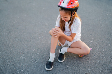 A girl wearing a helmet crying while touching her knee after falling off a skateboard on an asphalt road. © sementsova321