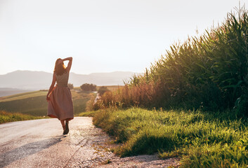 Confident young woman in stylish beige dress walking along asphalt road during warm golden hour light, holding skirt gracefully while enjoying peaceful countryside landscape and soft summer breeze