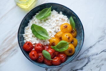 Cottage cheese caprese with mixed roasted tomatoes and green basil in a blue bowl, horizontal shot on a white granite background, high angle view
