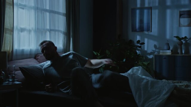 Full shot of young man suffering from PTSD twisting and jerking in bed while experiencing nightmare in dream, waking up, jumping off bed, sitting up in panic, rubbing face and drinking glass of water