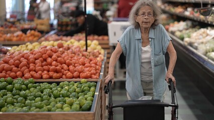An elderly woman navigates through a supermarket with her walker, carefully selecting fresh vegetables among vibrant displays of produce.