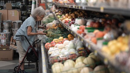 Disabled elderly woman navigates a grocery store with her walker while selecting fresh fruits and vegetables from the shelves during daylight hours.