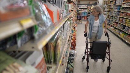 An elderly woman with a walker navigates the aisles of a grocery store, carefully selecting items from the shelves during her shopping trip.