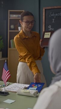 Vertical medium shot of young Caucasian female teacher using sticky notes on classroom objects while practicing vocabulary with students during interactive lesson in multicultural class