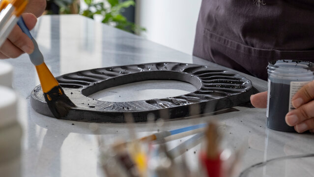 A close-up shot of a beautiful middle-aged woman painting a clay ceramic frame black at a table in an art studio, with artistic paintings adorning the background - Powered by Adobe