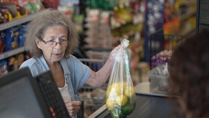 An elderly woman with gray hair is grocery shopping, holding a bag of lemons. She engages with the cashier in a bustling store setting filled with various food items.